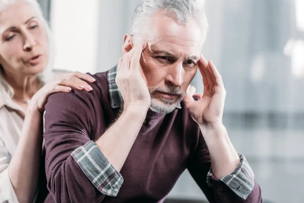 A man with gray hair holding his head in discomfort while a woman with gray hair offers support from behind.